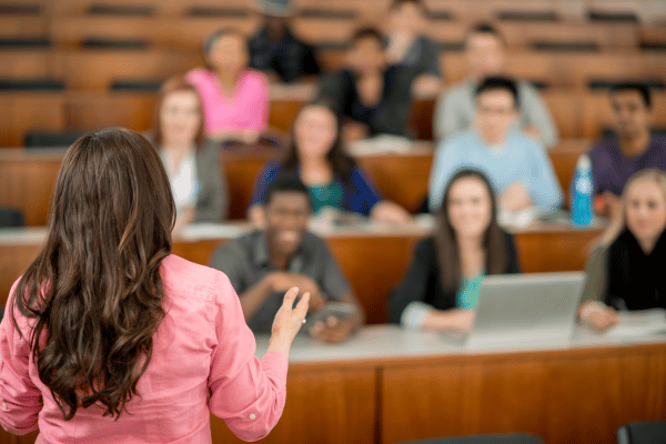 Professora de costas para câmera observa estudantes ao fundo, em plateia