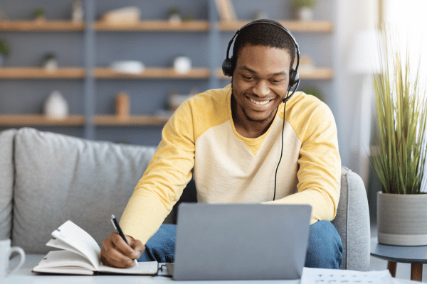 Estudante sorri para a tela do computador enquanto anota em seu caderno