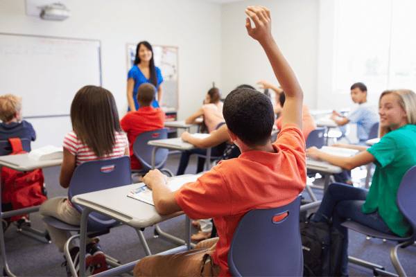 jovem levanta a mão em sala de aula para a professora ao fundo