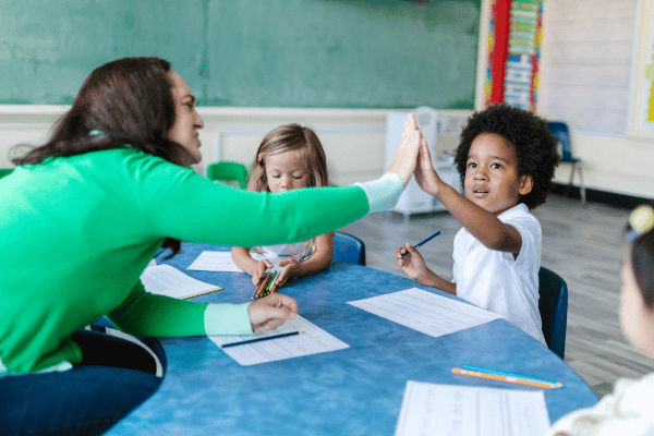 Pequenos estudantes na mesa, um aperta a mão de uma professora